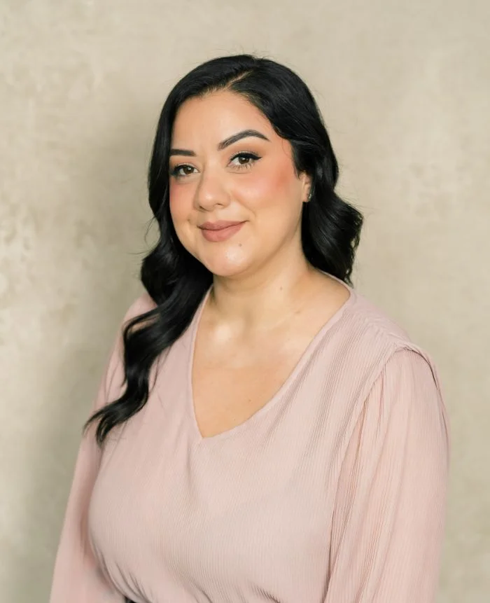 Portrait of a woman with black wavy hair wearing a light pink blouse against a beige background.