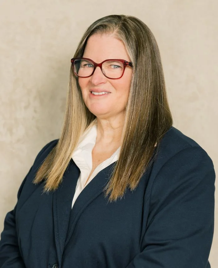 Professional portrait of a woman with long straight hair, red glasses, white blouse, and navy blazer.