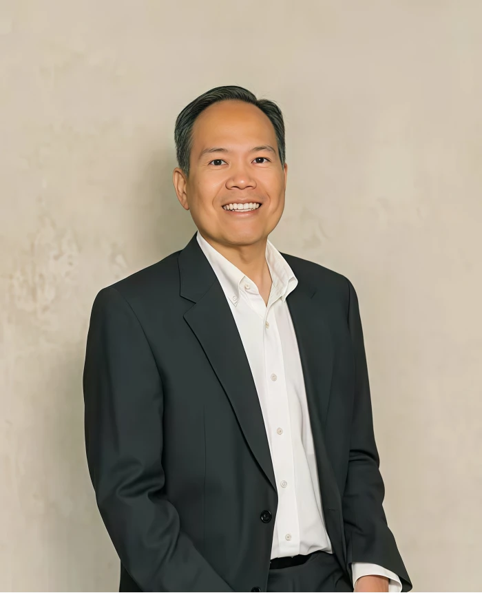 Professional headshot of a smiling man in a black suit and white shirt, beige background.