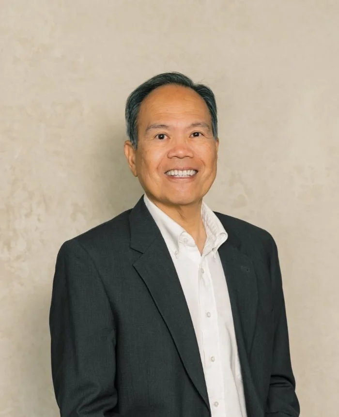 Professional headshot of a middle-aged man wearing a dark suit jacket and white shirt, smiling against a beige background.