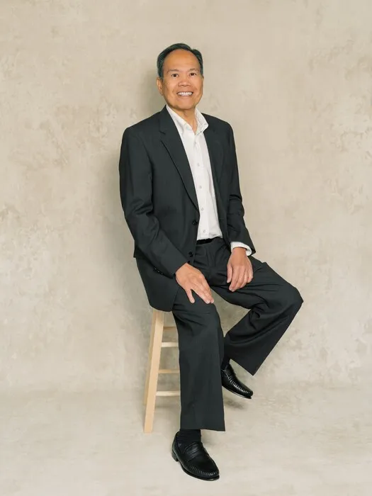 Middle-aged man in black suit and white shirt sitting on wooden stool against neutral background.