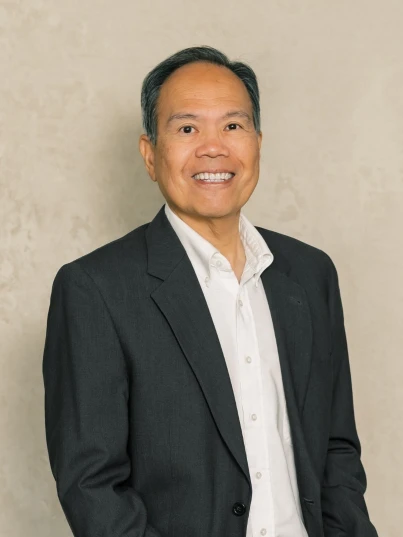 Portrait of a middle-aged man in a dark suit jacket and white dress shirt, smiling against a beige background.