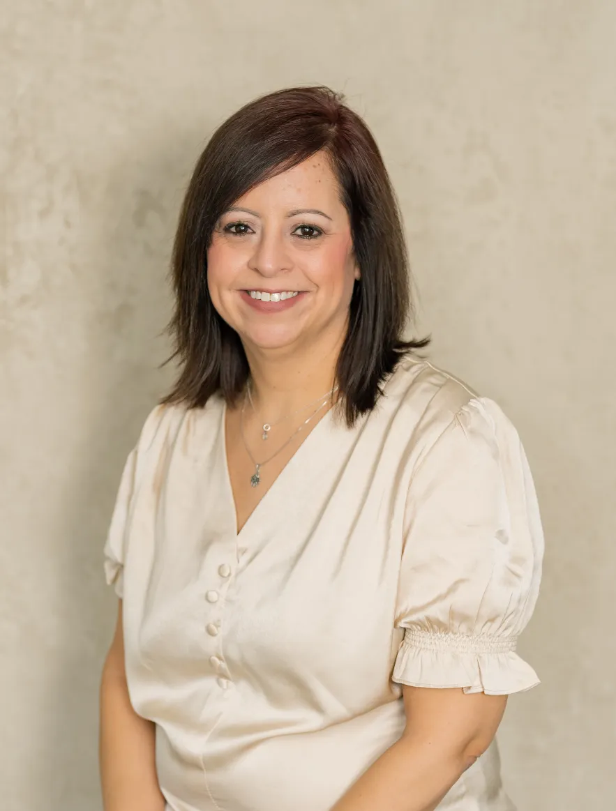 Portrait of a smiling woman with dark hair wearing a cream satin blouse and necklaces.