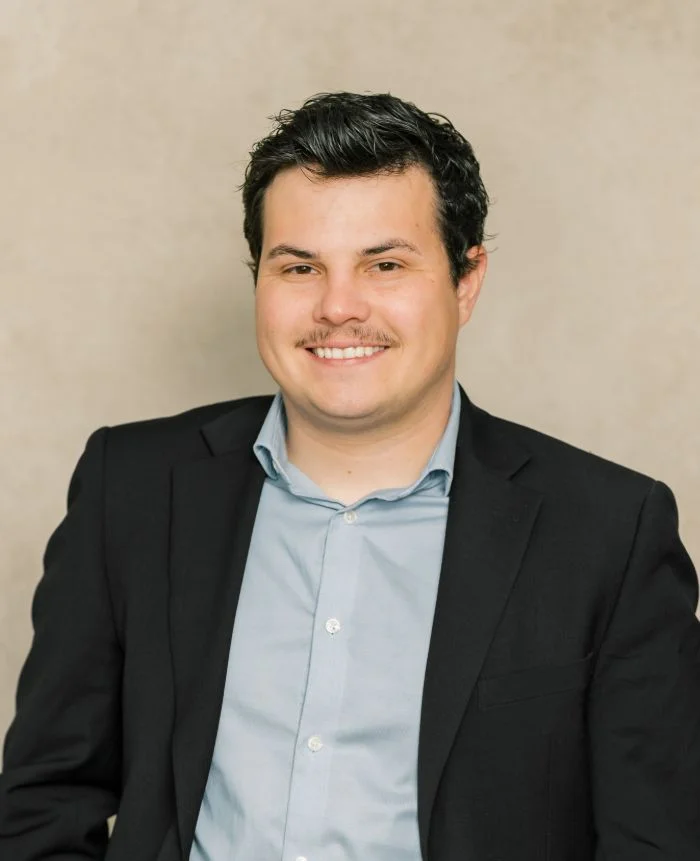 Professional portrait of a smiling man with dark hair, wearing a black blazer and light blue shirt.