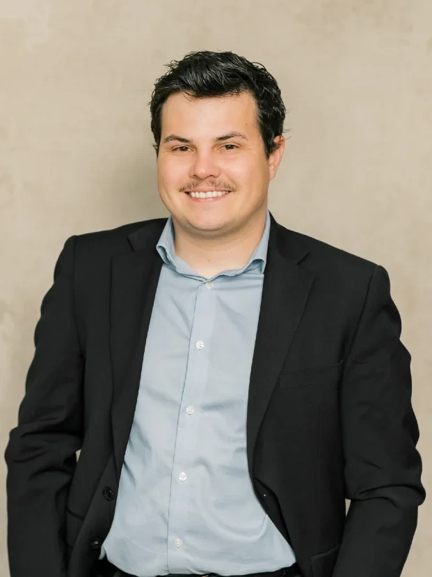 Professional portrait of a smiling man with dark hair, wearing a black blazer and light blue shirt.
