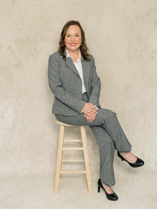 Professional woman in gray suit and black heels seated on wooden stool against beige background.
