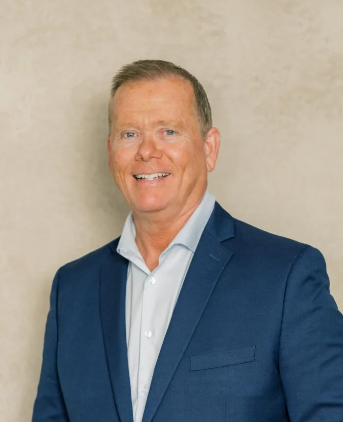 Professional portrait of a middle-aged man in a navy blue suit and light blue shirt against a beige background.