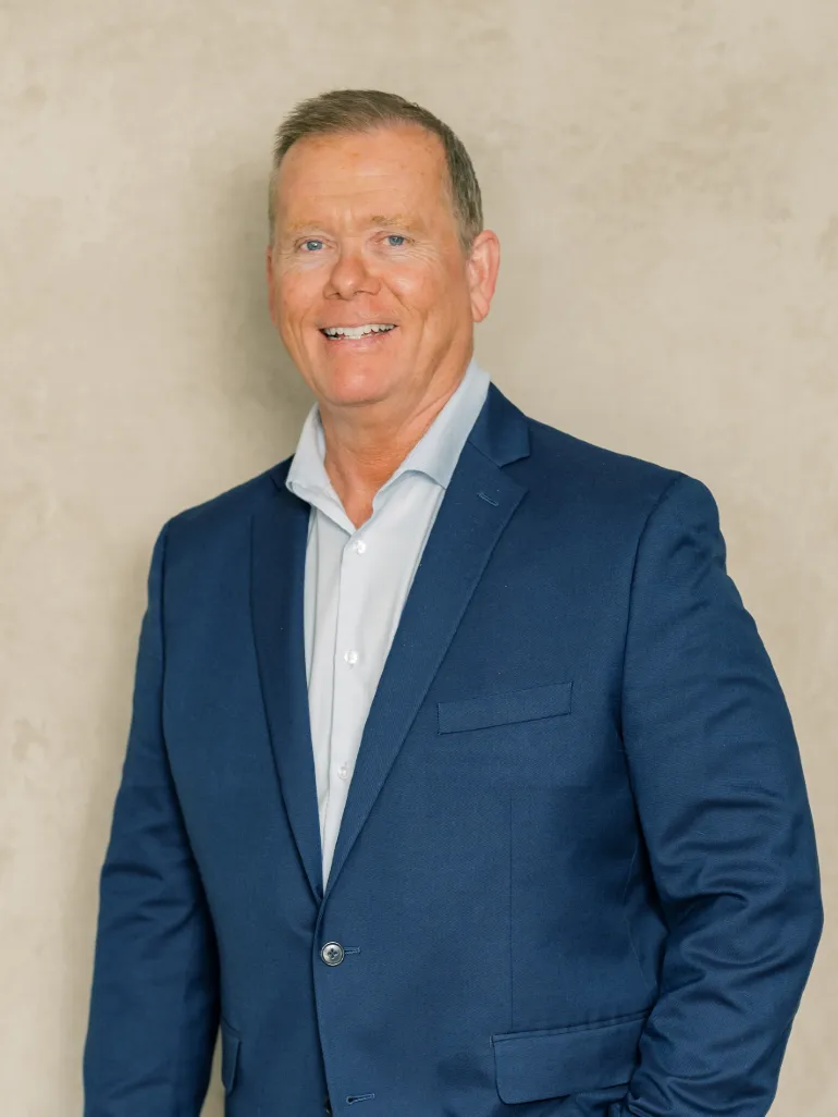 Professional middle-aged man wearing a blue suit and white shirt, smiling against a beige background.