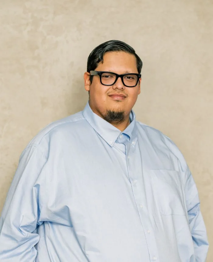 Portrait of a man with black glasses and neatly combed hair wearing a light blue button-up shirt.
