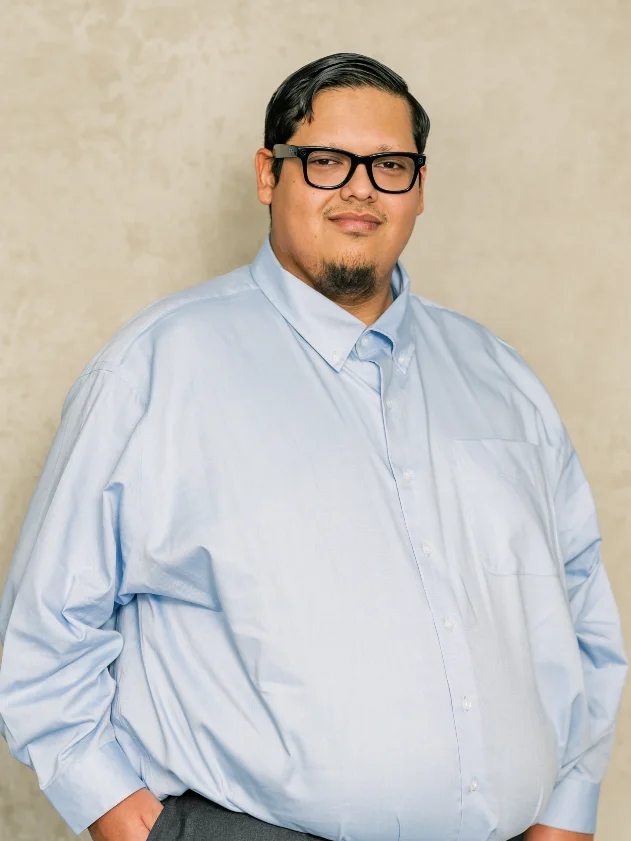 Portrait of a man wearing black glasses and a light blue button-up shirt against a beige background.