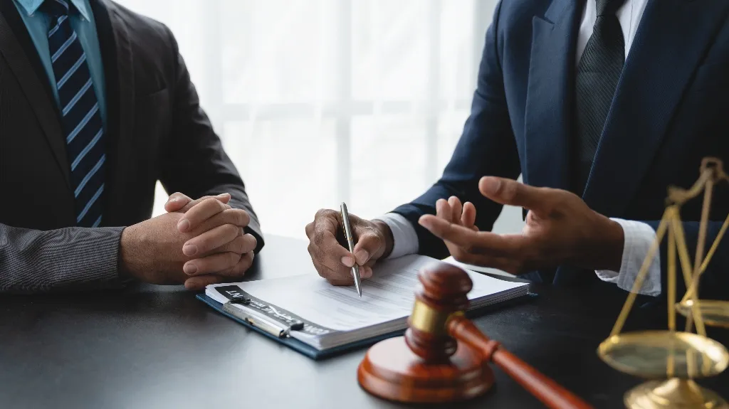 Two men in suits at a desk with legal documents, a gavel, and scales of justice.