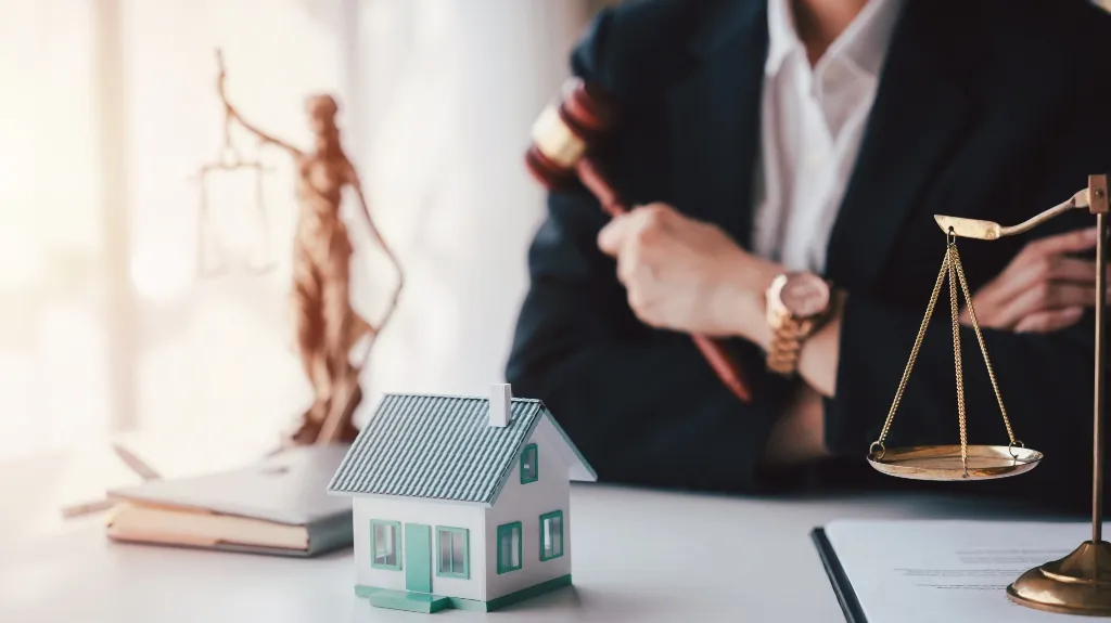 Person in formal attire holding a gavel, with house model, scales of justice, and law book on desk.