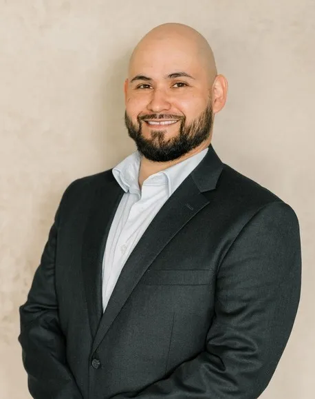 Professional portrait of a smiling bald man with a beard wearing a dark suit and light blue shirt.