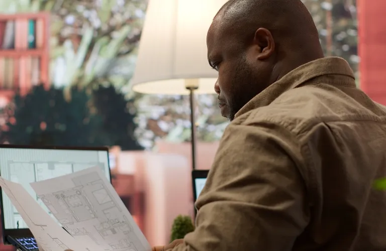 Man in brown shirt reviewing architectural blueprints with laptop and lamp in background.