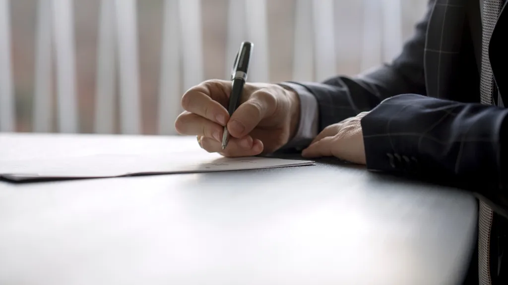 Close-up of a person in a dark suit signing a document on a white table.
