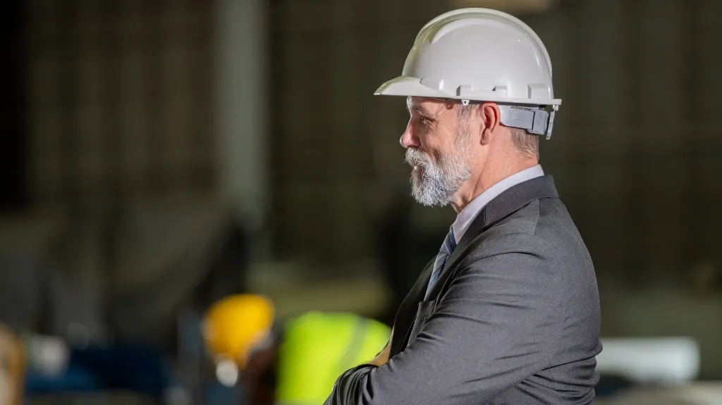 Middle-aged man with gray beard wearing white hard hat and suit in industrial setting.