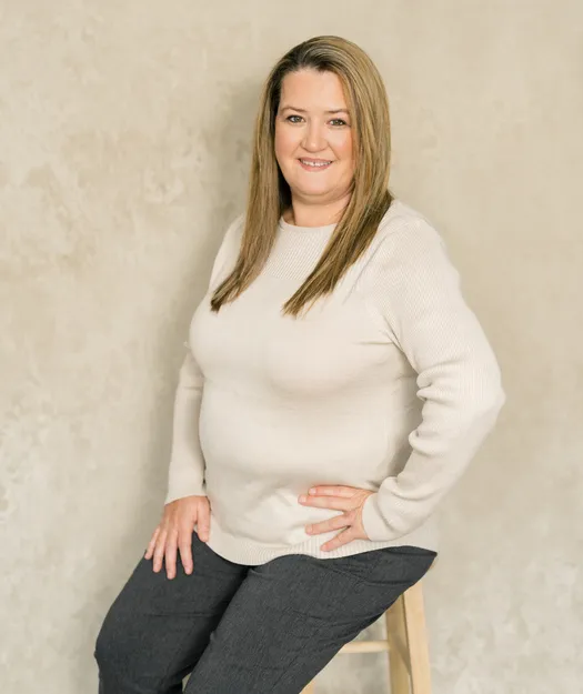 Professional portrait of a woman with long blonde hair, wearing a beige sweater and dark pants, seated on a wooden stool against a neutral background.