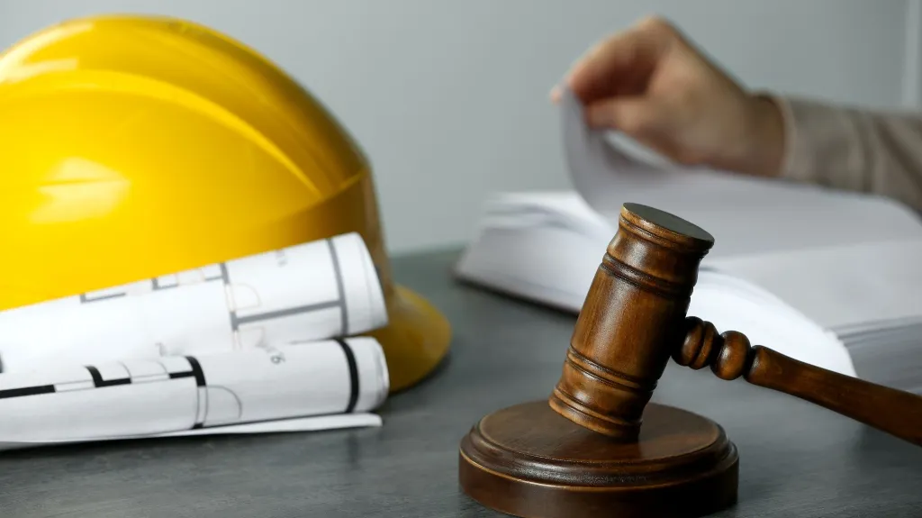 Wooden judge's gavel, yellow construction helmet, and rolled architectural blueprints on table.