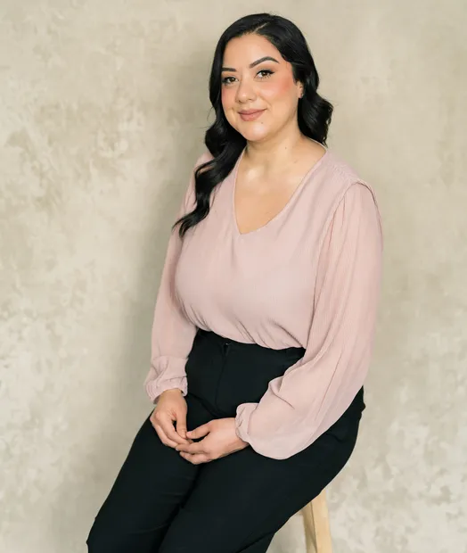 Portrait of a woman with dark hair, wearing a light pink blouse and black pants, seated on a stool.