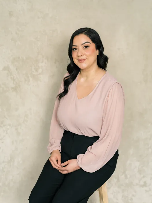 Professional portrait of a woman with long dark hair, wearing a light pink blouse and black pants, seated against a neutral background.