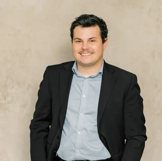 Portrait of a smiling man with dark hair wearing a black suit and light blue shirt.