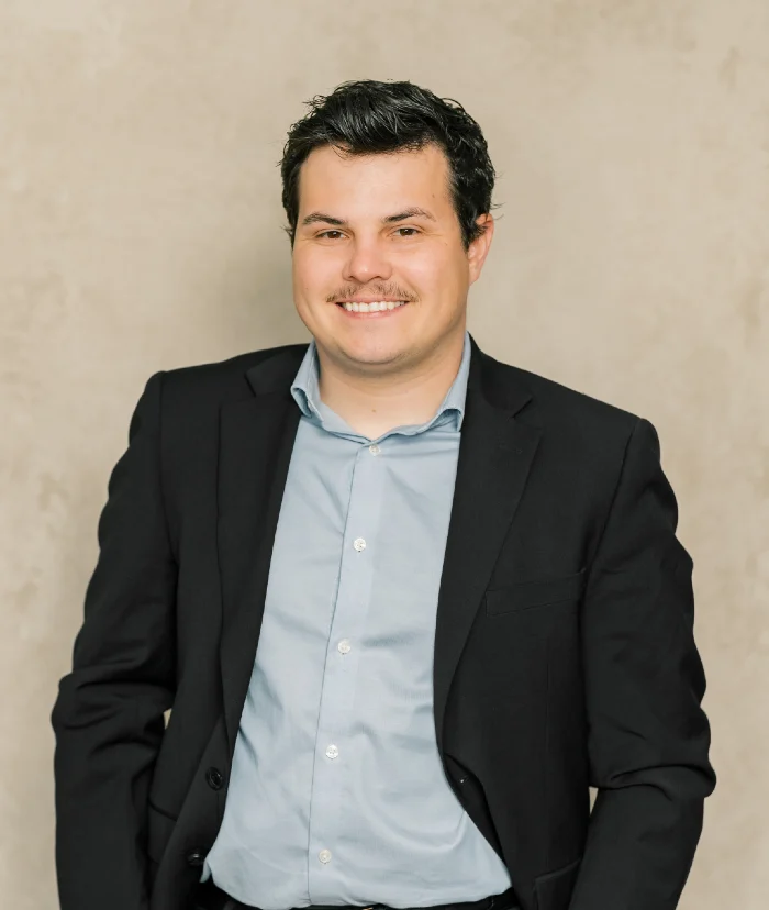 Professional portrait of a smiling man with dark hair wearing a black suit and light blue shirt against a beige background.