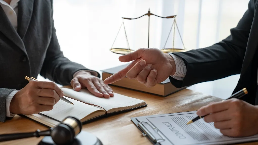 Two professionals in suits discussing a contract with legal scales and gavel on desk.