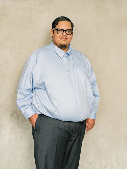Man with black hair and glasses wearing a light blue dress shirt and dark gray pants.