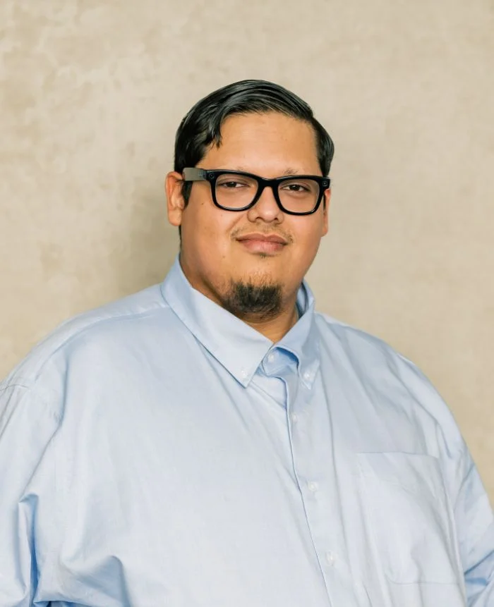 Portrait of a man wearing black glasses and a light blue button-up shirt against a beige background.