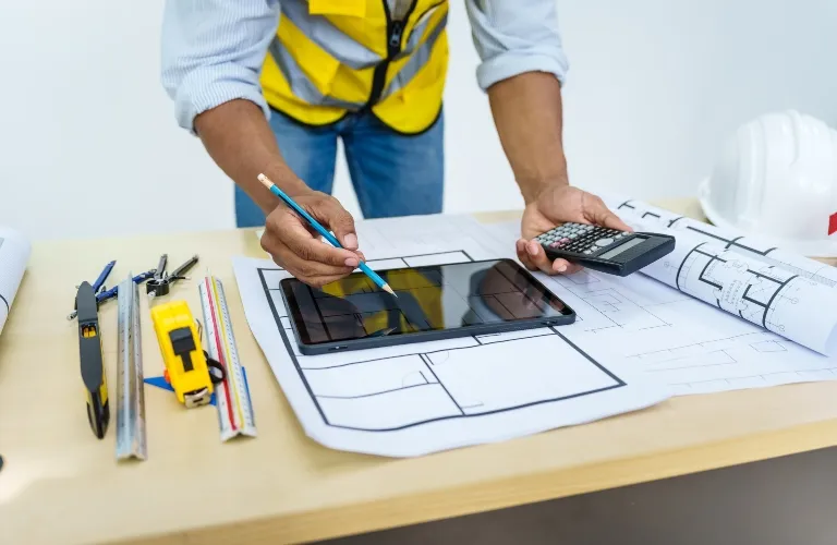 Architect in yellow safety vest using a tablet and calculator over blueprints on a desk.