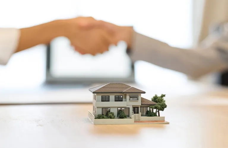 Miniature model of a two-story house on table with blurred handshake in background.