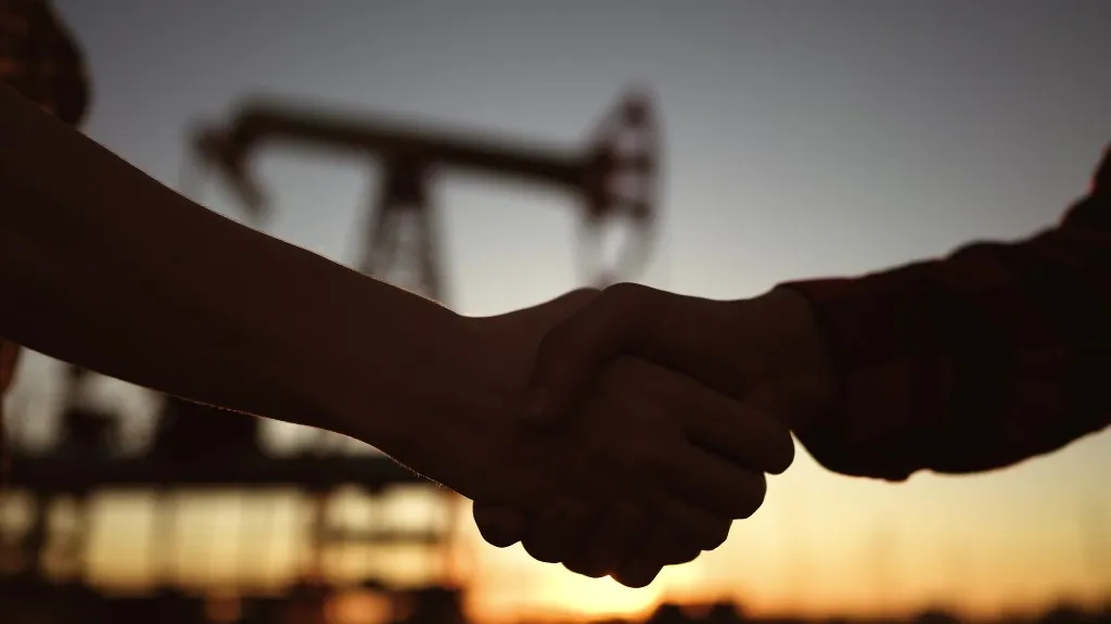 Two individuals shaking hands with an oil pumpjack silhouetted against a sunset background.