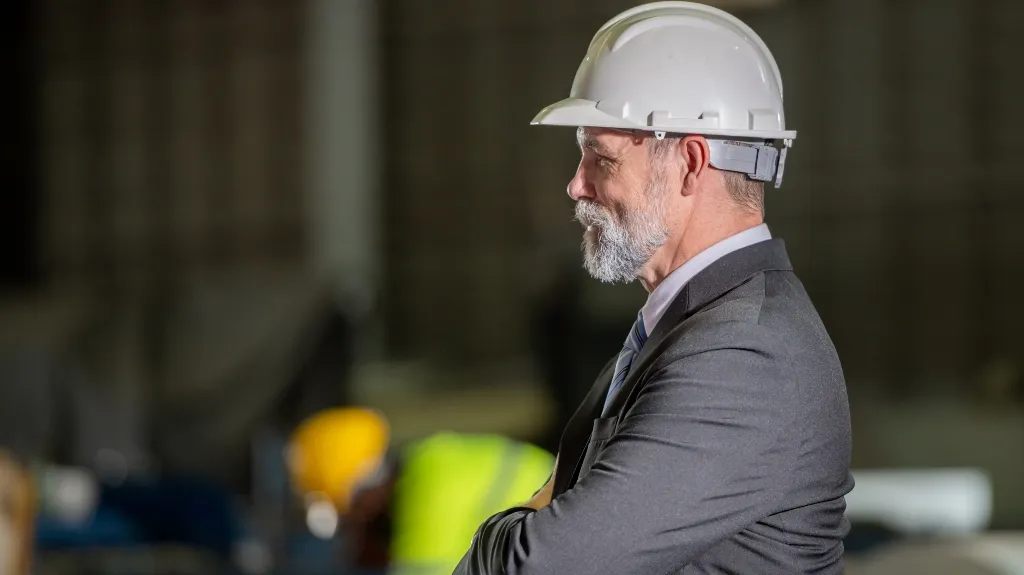 Middle-aged man in a suit and white hard hat standing with arms crossed in industrial setting.