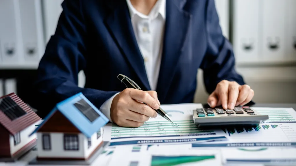 Person in business attire using a calculator and pen, analyzing financial documents with house models nearby.