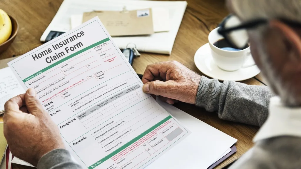 Elderly person reviewing a home insurance claim form at a wooden table with coffee.