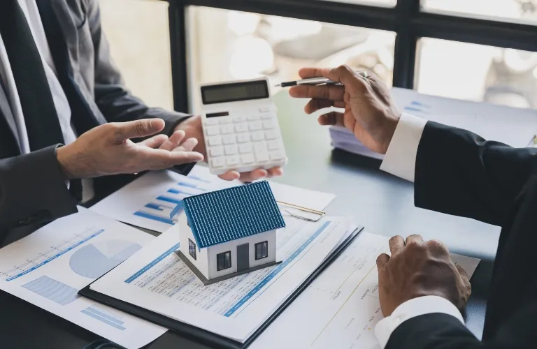 Two professionals in suits discussing financial documents with a small house model and calculator on the table.