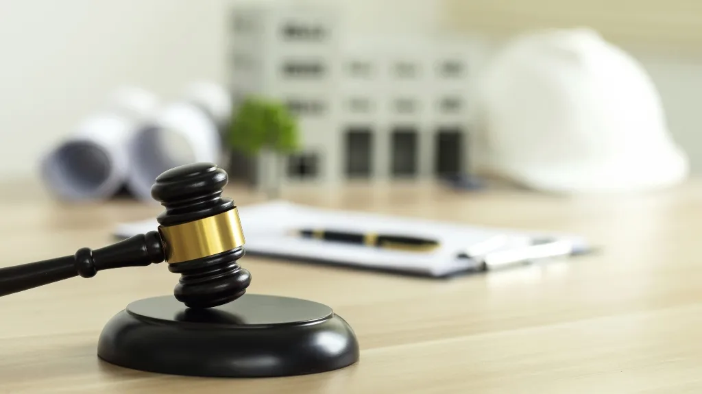 Wooden judge's gavel with gold band on desk, blurred construction helmet and blueprints in background.